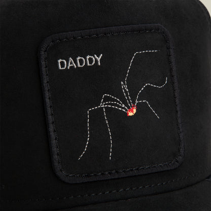 A black fabric patch from Goorin Bros., named The Suede Spider, featuring the word “DADDY” and an embroidered arachnid with a red spot, is sewn onto a black surface.