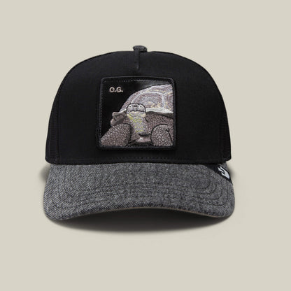 The Farm's "The OG Tortoise" is a black and grey denim trucker hat with a patch featuring an embroidered tortoise and "O.G." letters, crafted from durable denim for lasting style.