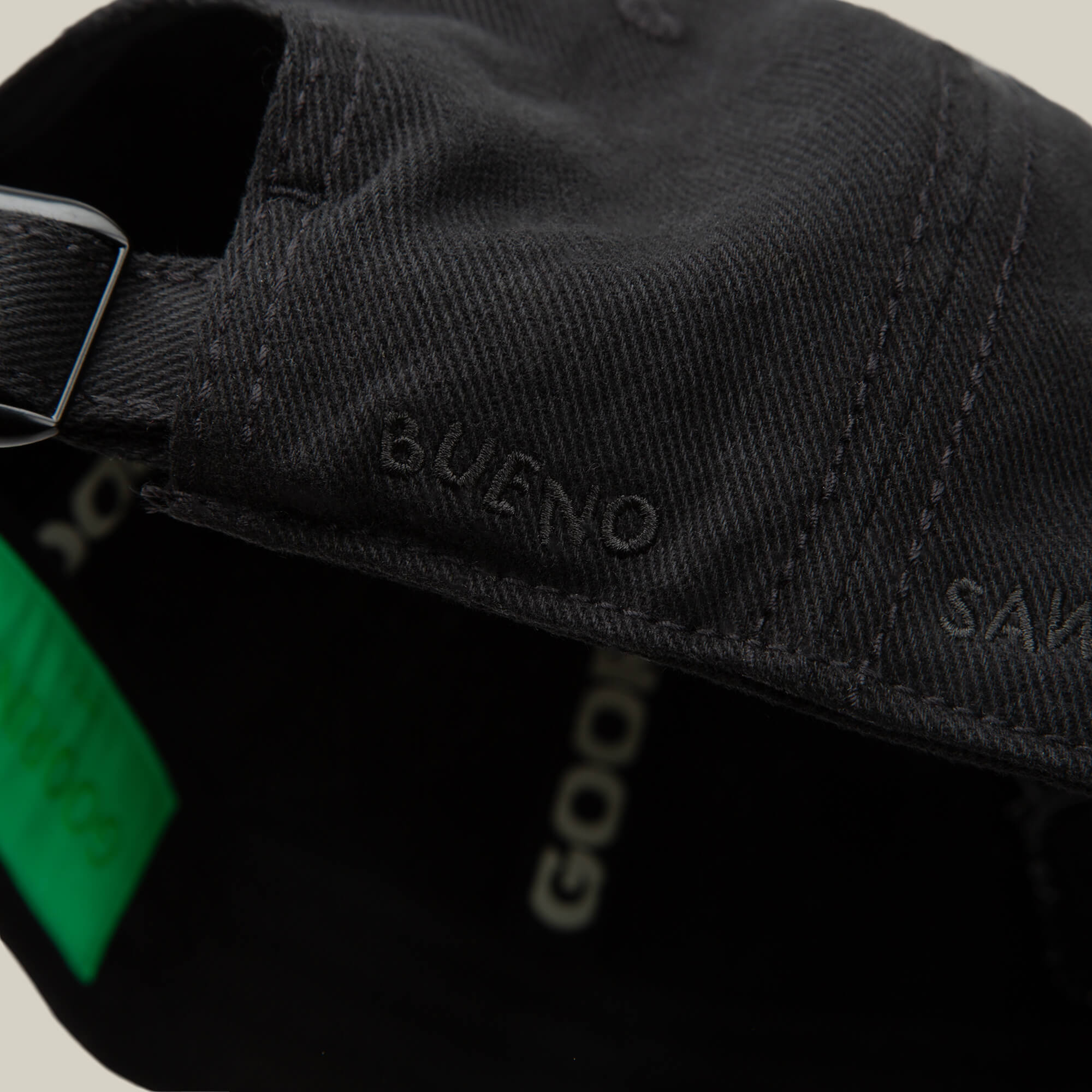 Close-up of the back of a black Fine Headwear cap from Goorin Bros., featuring "BUENO" embroidered near the adjustable strap for a stylish touch.