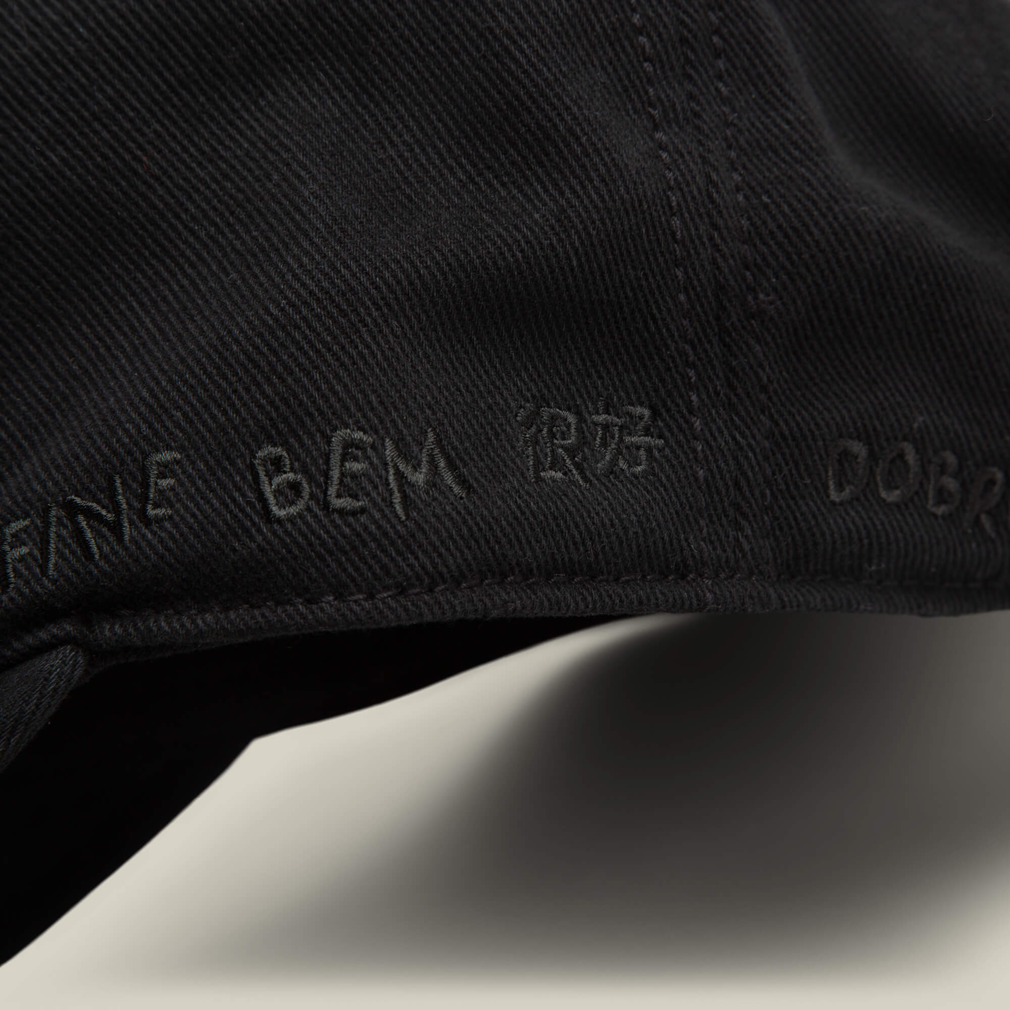 Close-up of black Fine Headwear by Goorin Bros., showcasing various embroidered words like "FANE BEM," "DOBR," a non-Latin symbol, and playful icons, including designs reminiscent of a smiley face.