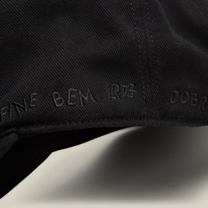 Close-up of black Fine Headwear by Goorin Bros., showcasing various embroidered words like "FANE BEM," "DOBR," a non-Latin symbol, and playful icons, including designs reminiscent of a smiley face.