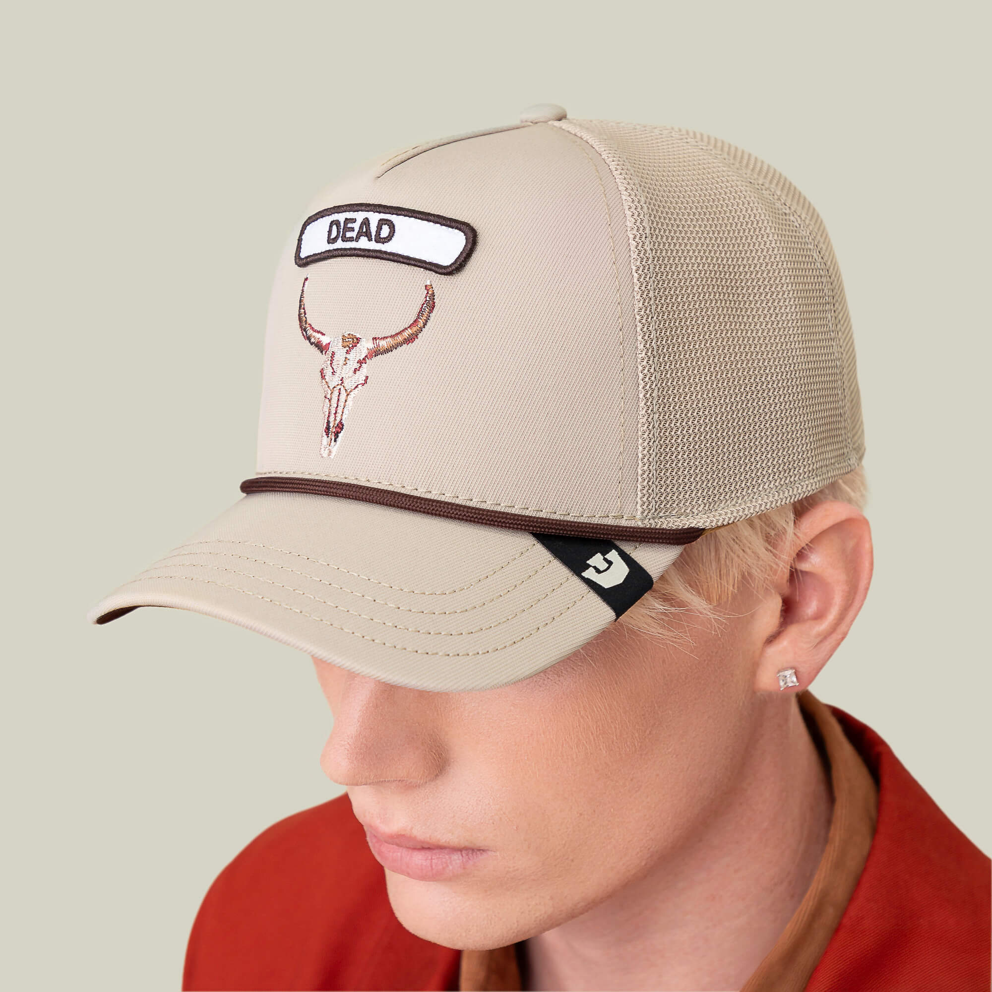 A person with light hair wears the Goorin Bros. GB2 Skull beige mesh trucker cap featuring a bull skull patch and "DEAD" embroidered on the front, paired with a rust-colored shirt that highlights desert-inspired tones.