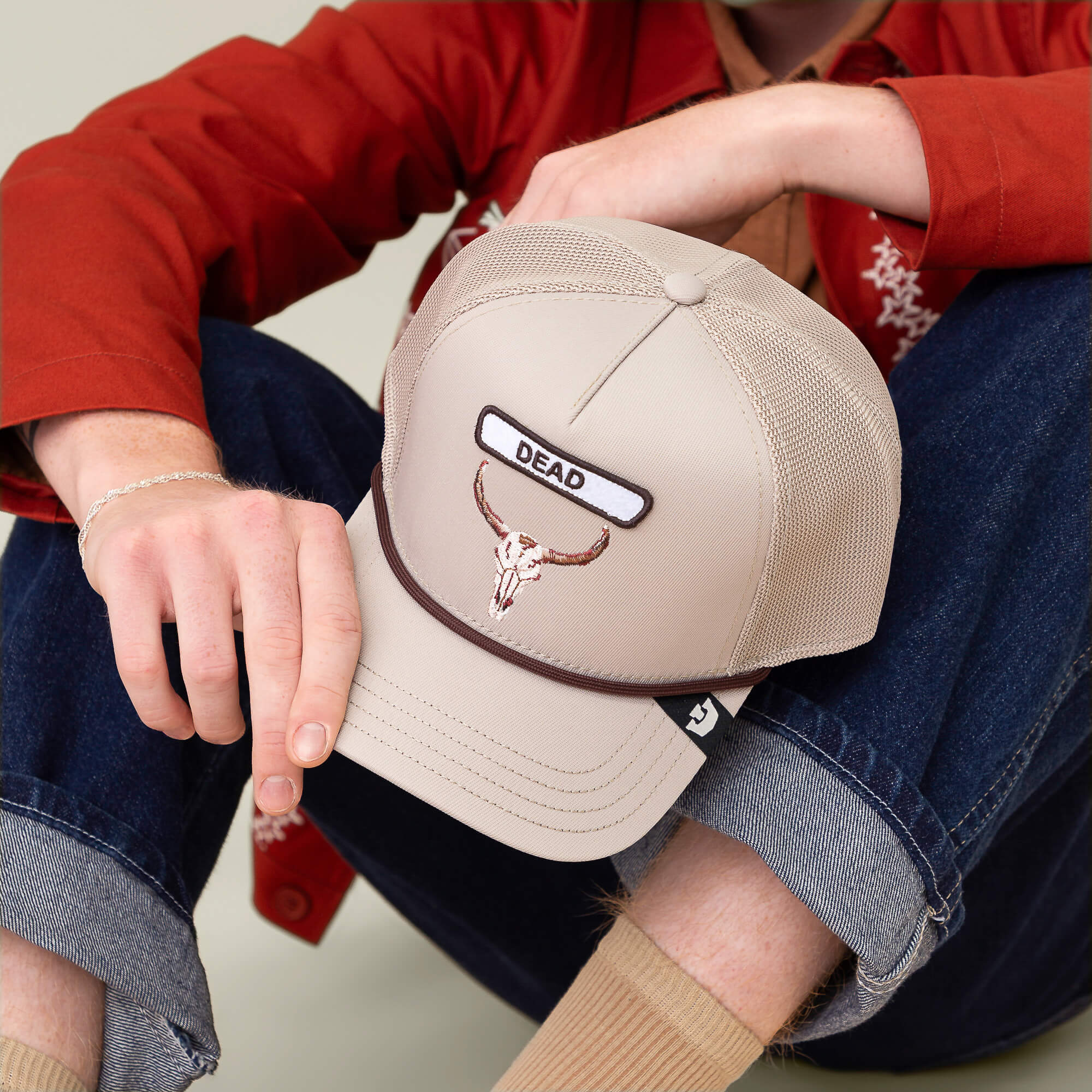 Someone in rolled-up jeans and a red jacket holds the Goorin Bros. GB2 Skull trucker hat, which features a skull patch and "DEAD" embroidered on the front in rugged desert tones.