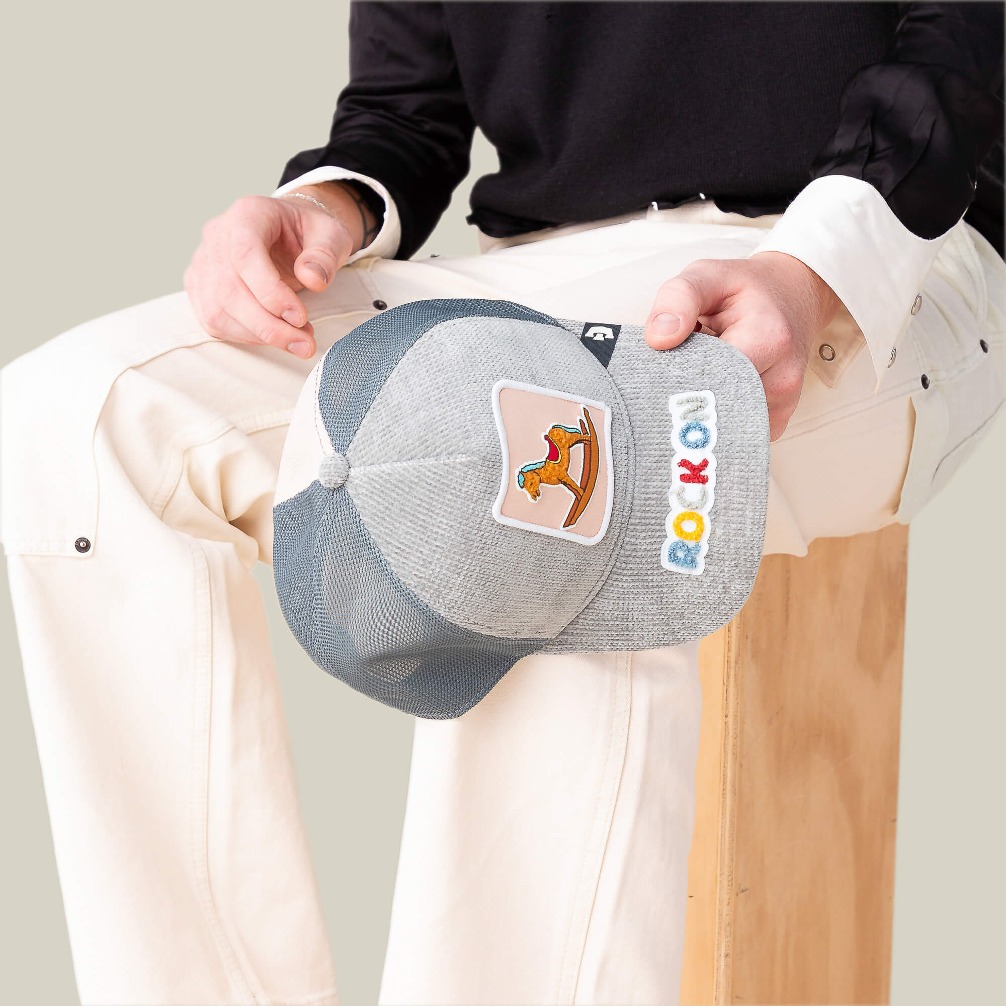 A person sits on a wooden block, holding the Goorin Bros. "Hobby Horse"—a gray mesh baseball cap featuring a rocking horse patch and colorful letters on the brim for a nostalgic streetwear vibe.