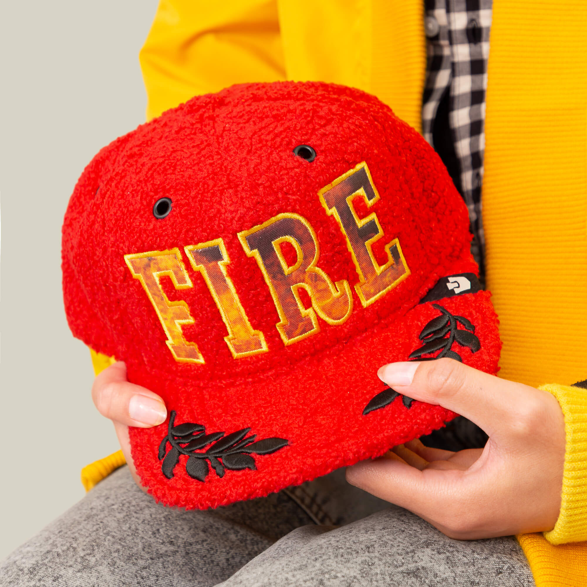 A person holds the Goorin Bros. Hot Fire hat—a bold red sherpa cap with "FIRE" in yellow letters and black leaf designs—while wearing a yellow jacket and plaid shirt.