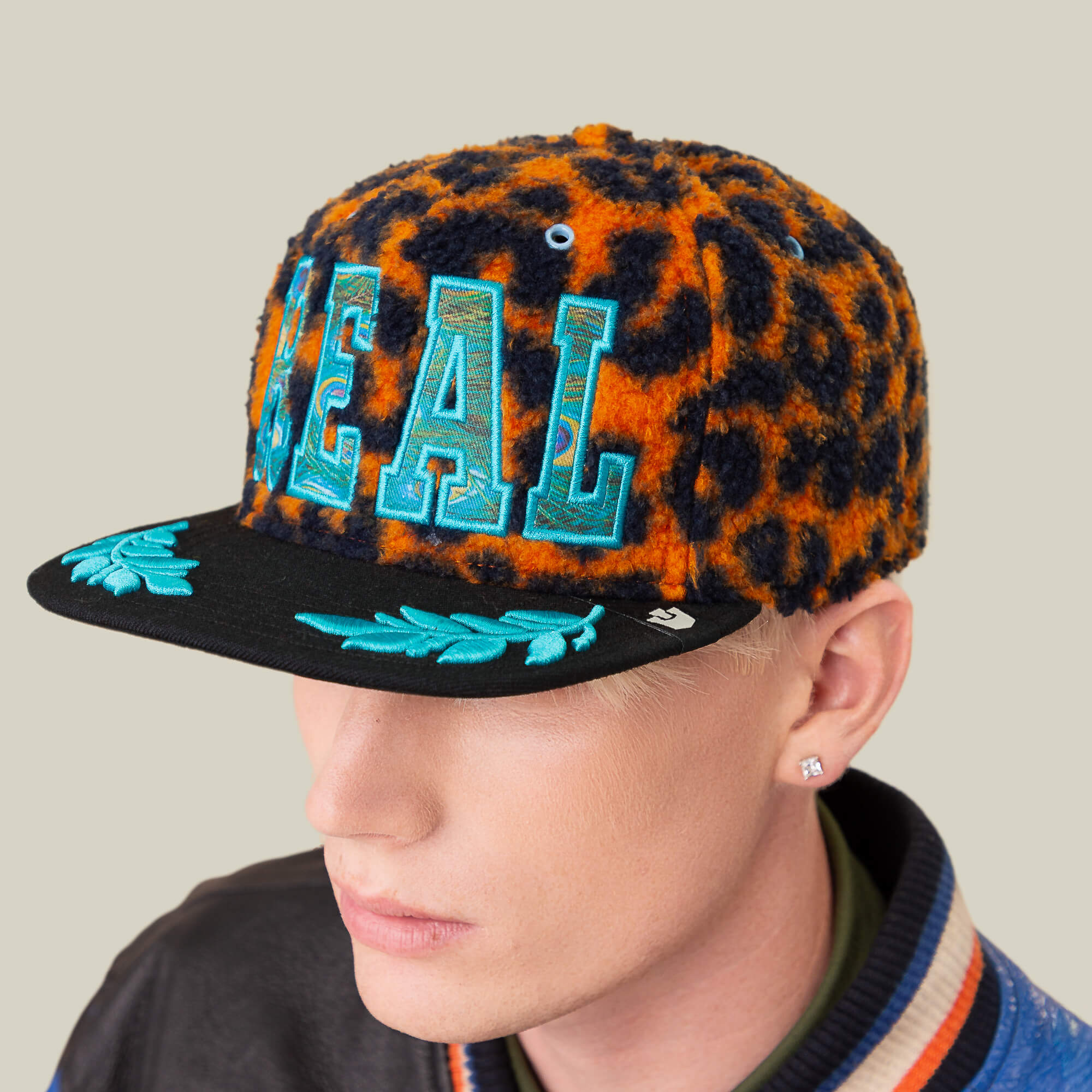 A person wearing the Goorin Bros. El Camino fuzzy orange leopard sherpa baseball cap with 'REAL' in blue letters and aqua embroidery on the brim, featuring a wider profile.