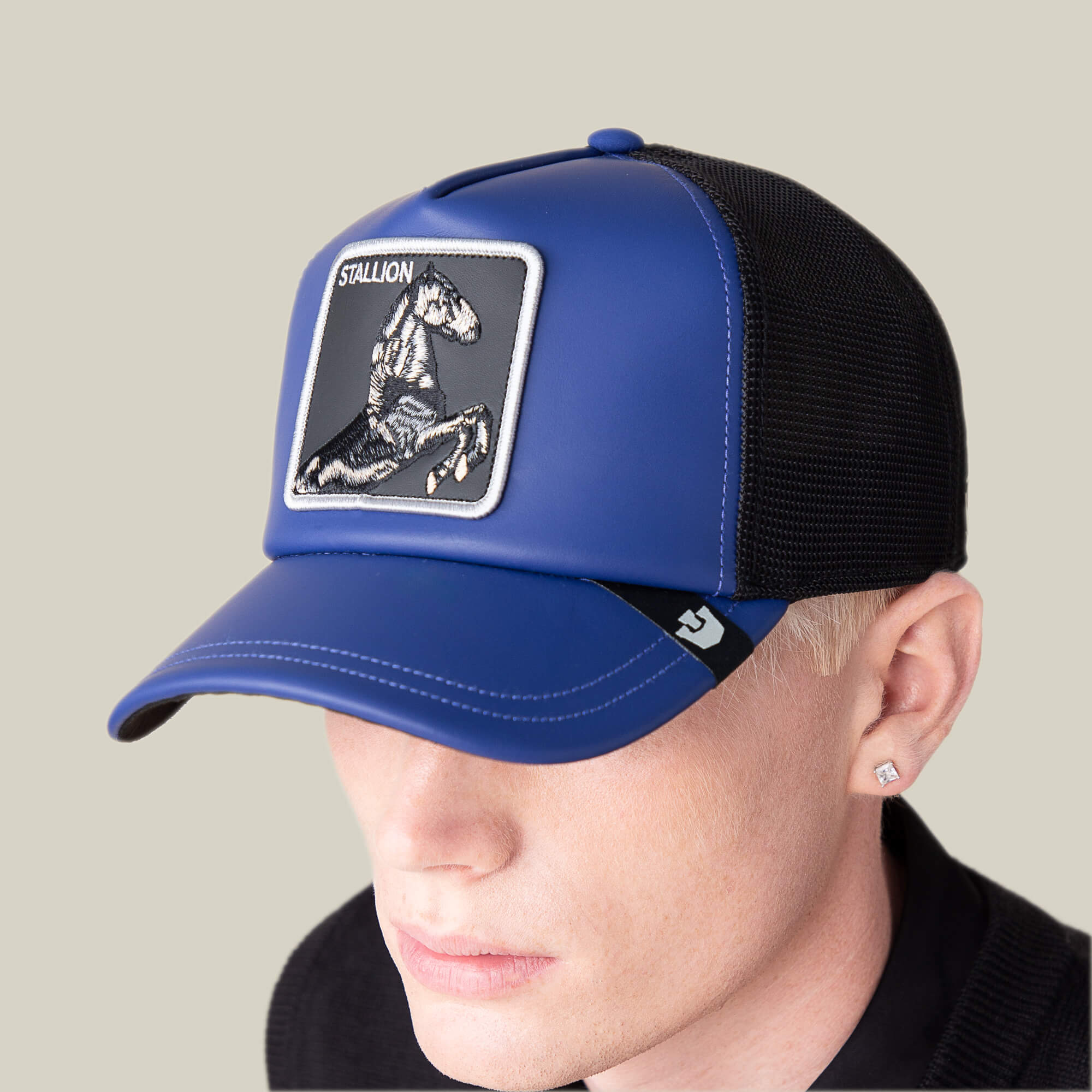 Wearing the Goorin Bros. LFG Stallion trucker hat—blue and black mesh with royal blue leather accents and a horse patch—this blond-haired, square-stud-wearing individual channels pre-game stallion style.