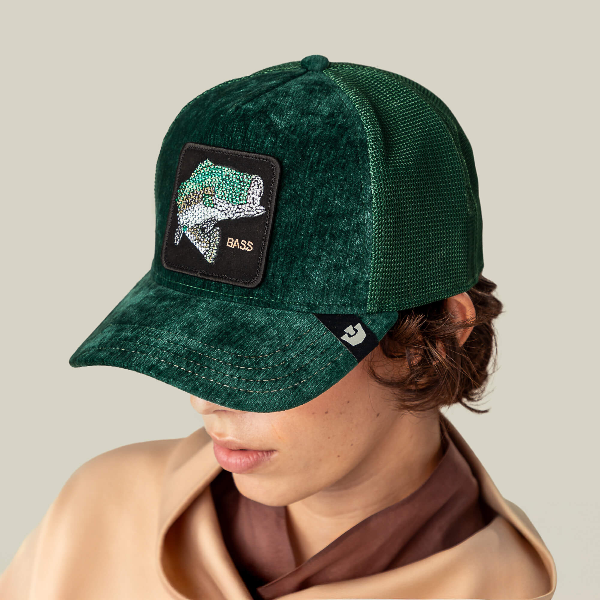 A person wears the Goorin Bros. Iced Bass green mesh trucker hat, featuring a fish patch with "BASS," adding a luxurious touch as they look down against a plain background.