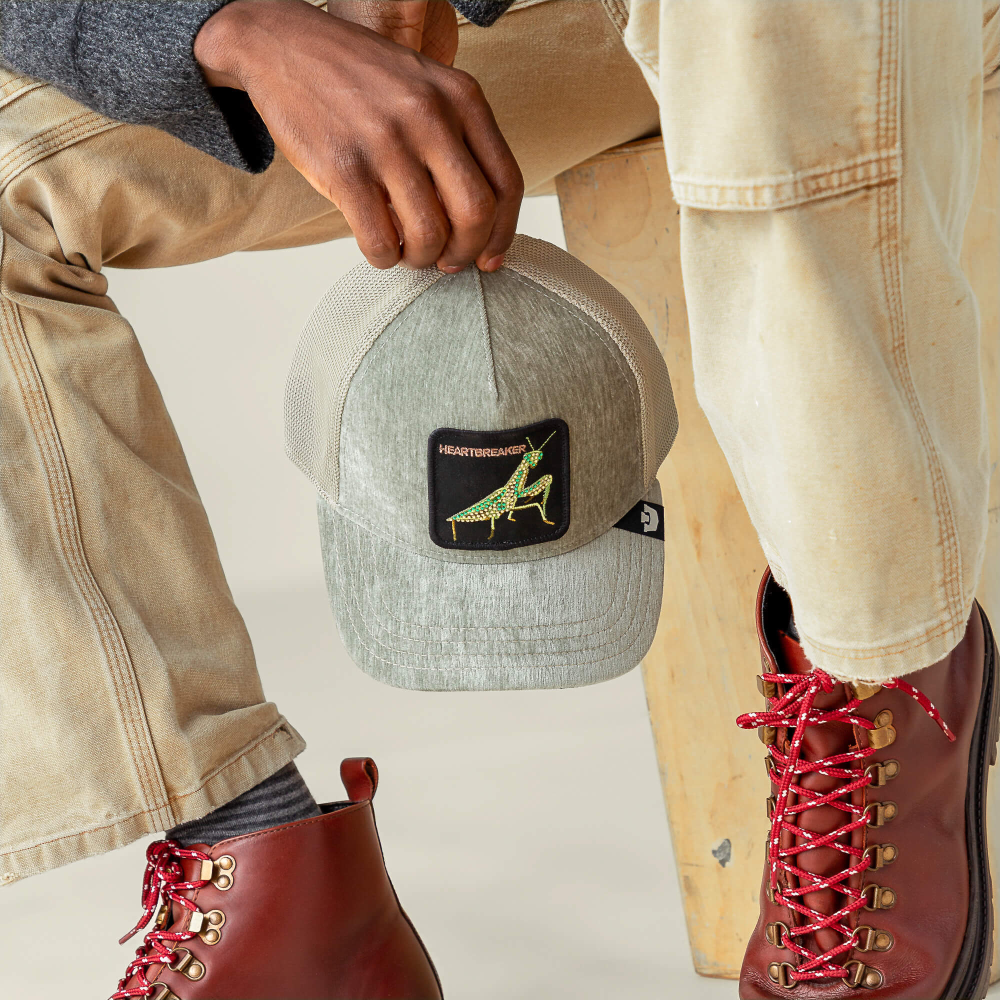 Sitting on a wooden stool, a person holds the Goorin Bros. Iced Mantis gray trucker hat with a dinosaur patch; beige pants and red lace-up boots complement champagne mesh under a sequined, armor-inspired jacket.