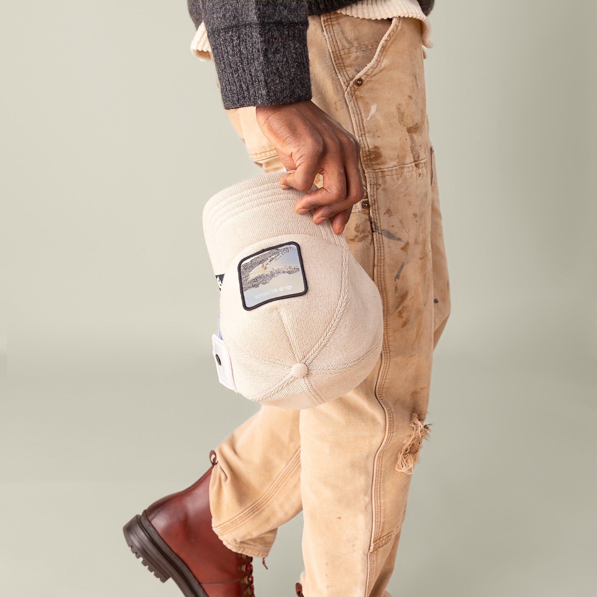 A person in tan, paint-splattered pants and brown boots holds the beige AllmiMator cap by Goorin Bros., shown waist-down against a plain background for a classic, vintage-inspired look.