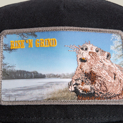 A close-up of the Goorin Bros. Morning Wood cap’s black crown displays an embroidered beaver and the text RISE ‘N GRIND, set against a snowy landscape.