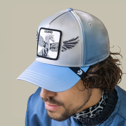 Man wearing the Goorin Bros. Flare Up cap, a light blue and gray hat featuring a winged Pegasus patch labeled Legend, paired with a blue jacket with contrast stitching over a black and white patterned shirt.