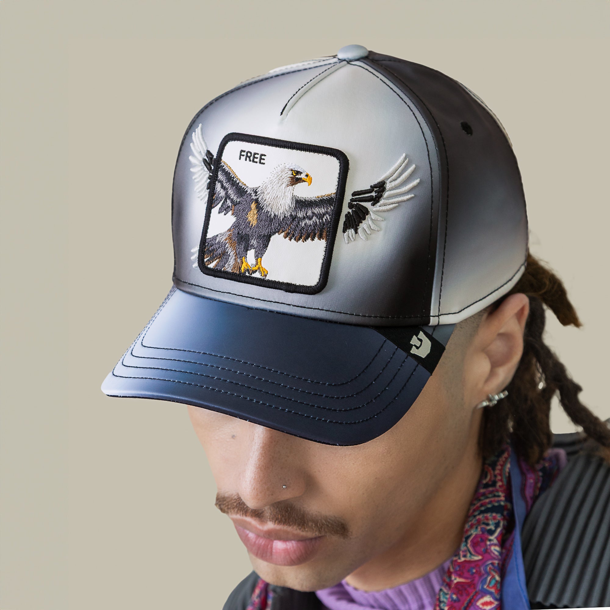 A person wears the Goorin Bros. "Steal This Hat," featuring a black-and-grey gradient, a metallic finish, a bold bald eagle patch, and an embroidered “FREE” patch on the front.