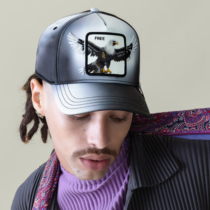 Someone sporting the Goorin Bros. "Steal This Hat," a black-and-grey gradient cap featuring a bold bald eagle patch and "FREE" on the front, paired with a purple turtleneck, pinstriped jacket, and patterned scarf.