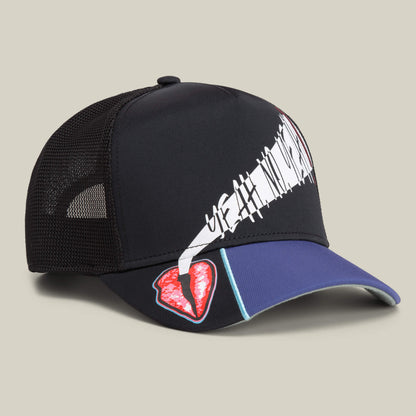The Means Yes hat by Goorin Bros. features black mesh, a white abstract graphic on the front, and a red broken heart patch on the blue brim—a bold accessory shown against a neutral background.