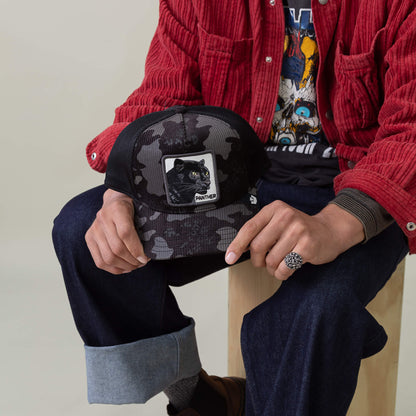 A person in a red jacket and jeans sits on a stool, holding the Goorin Bros. Panther Camo mesh cap featuring a panther patch on the front.