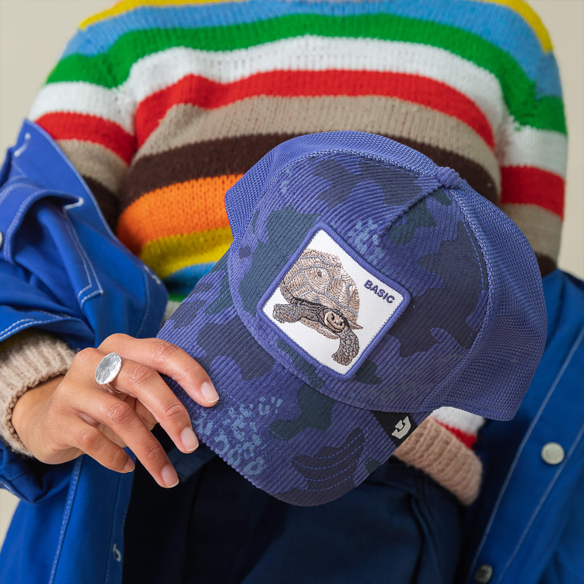 A person in a colorful striped sweater and blue jacket holds the Goorin Bros. Turtle Camo cap, featuring blue camo corduroy and an embroidered turtle patch that says BASIC.