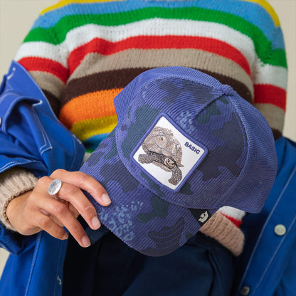A person in a colorful striped sweater and blue jacket holds the Goorin Bros. Turtle Camo cap, featuring blue camo corduroy and an embroidered turtle patch that says BASIC.