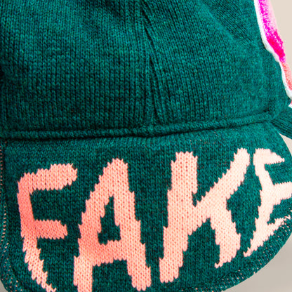 A close-up of the Goorin Bros. "F is for Fake" green knit fabric with FAKE stitched in large pink letters—a bold accessory for cold weather.