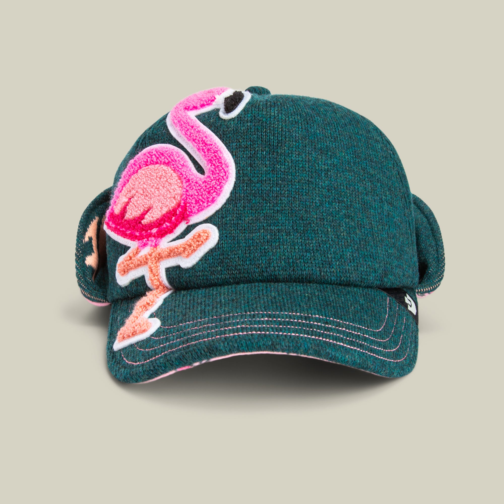The Goorin Bros. "F is for Fake" trucker baseball cap in GUARD features a green fabric with ear flaps and a large pink flamingo patch on the front, shown against a plain beige background.