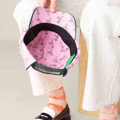 A person holds an open Goorin Bros. "F is for Fake" pink baseball cap featuring a flamingo pattern inside, styled with white pants, orange socks, and black loafers for a quirky cold-weather look.
