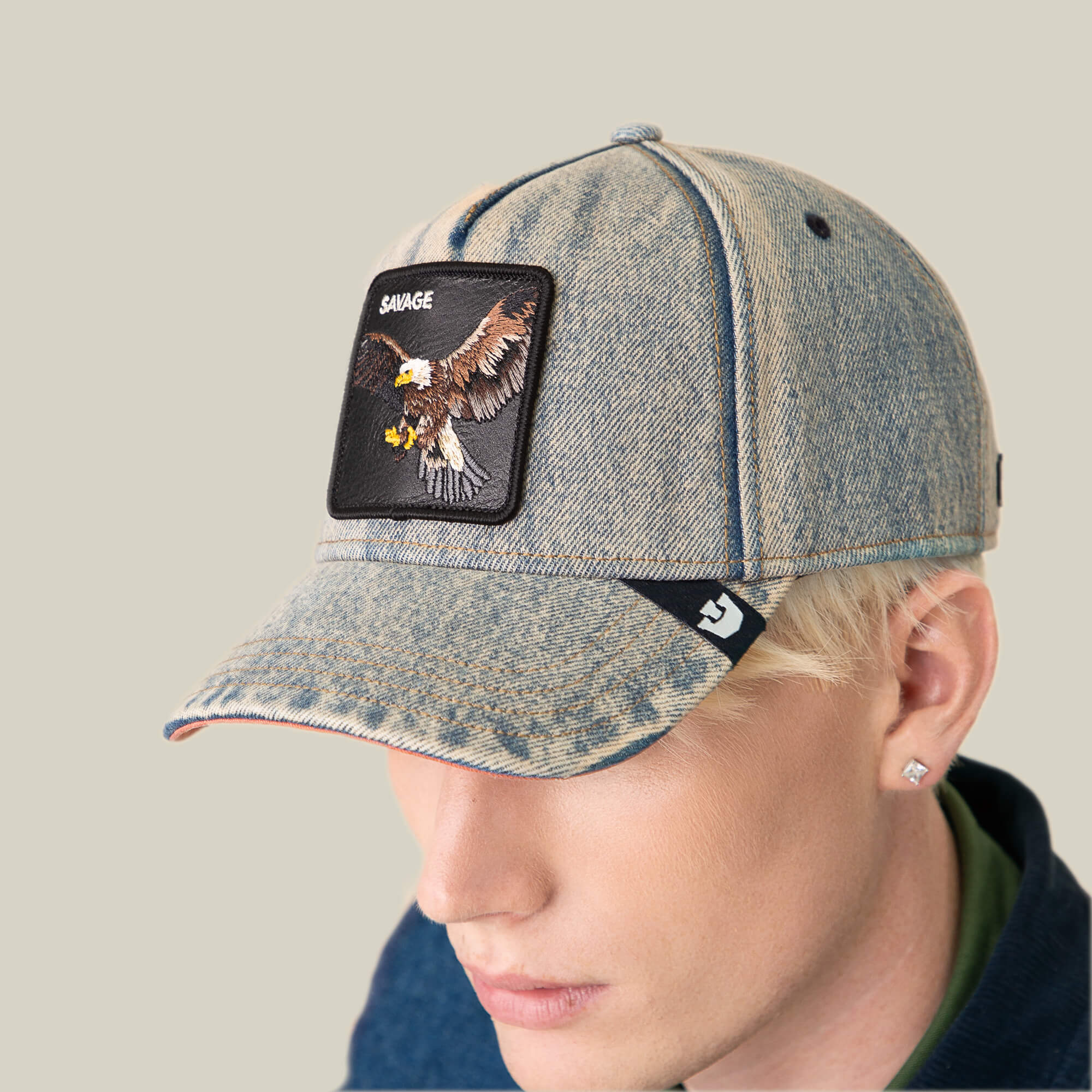 A person wears the Goorin Bros. Frequent Flyer cap—a light-wash denim hat featuring an eagle patch, “SAVAGE” label, visible seams, and a small logo tag that accent the classic vintage style.