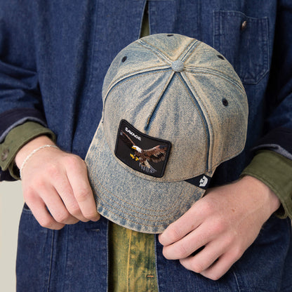 A person in a denim jacket and green shirt holds Goorin Bros.' Frequent Flyer cap, a vintage-style denim hat with an eagle patch and the word SAVAGE on the front.