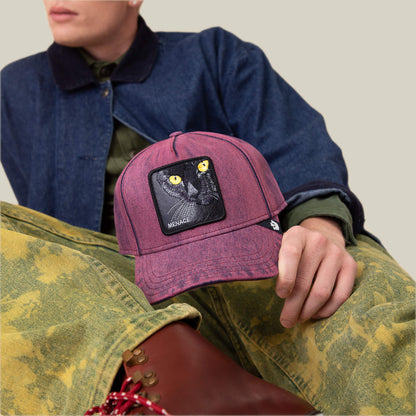 Wearing an overdyed denim jacket and yellow patterned pants, this person holds the Goorin Bros. Rail Bird cap, featuring a black cat patch and “MENACE” on the front, radiating pure troublemaker energy.
