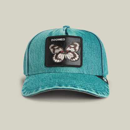 The Goorin Bros. Drawing Dead cap is a turquoise denim hat with thrift-store style, featuring a butterfly patch and "DOOMED" stitched above it.