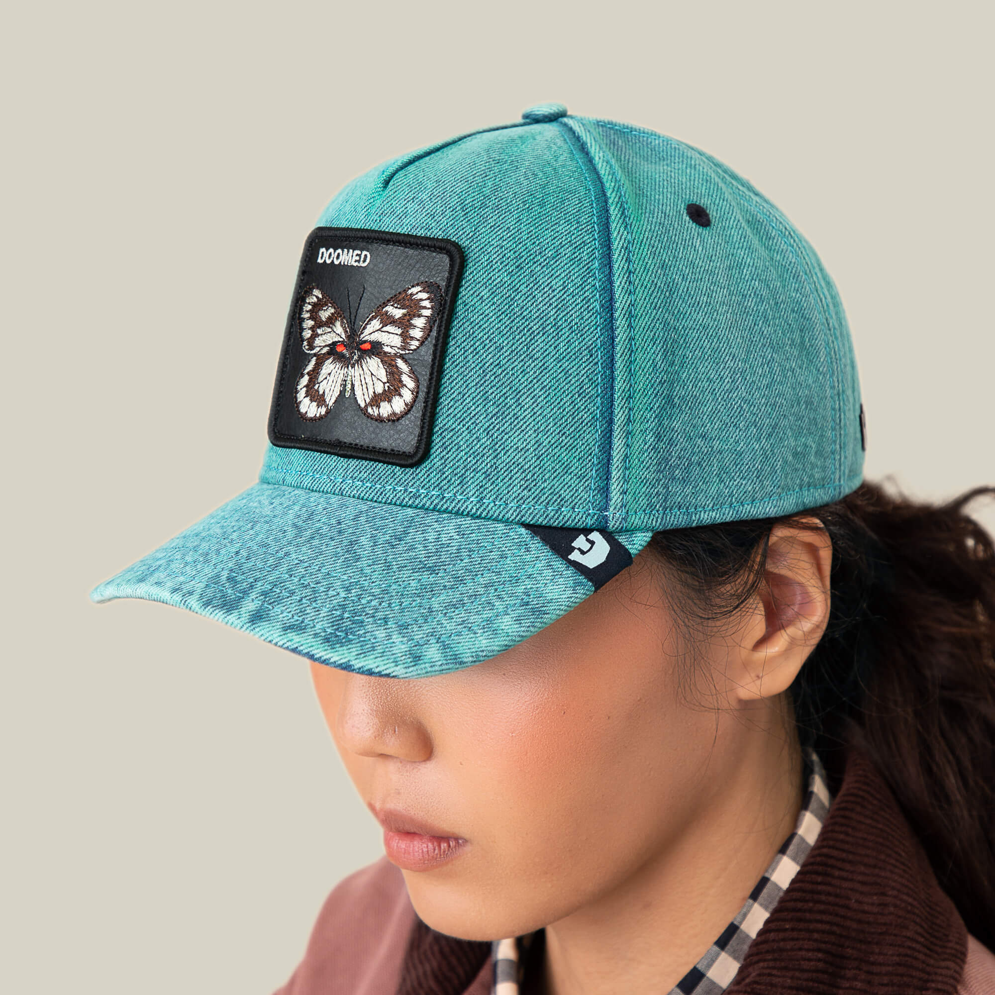 Person wearing the Goorin Bros. Drawing Dead denim hat with a butterfly patch that says DOOMED, paired with a plaid shirt, set against a plain beige background.