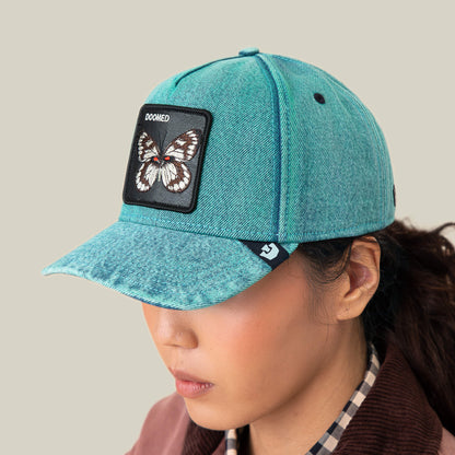 Person wearing the Goorin Bros. Drawing Dead denim hat with a butterfly patch that says DOOMED, paired with a plaid shirt, set against a plain beige background.