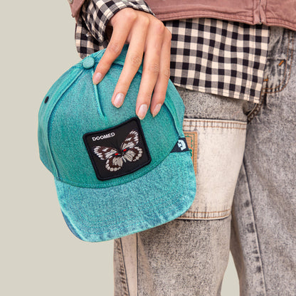 A person holds a Drawing Dead cap by Goorin Bros., sporting a checkered shirt and patchwork jeans for a cool thrift-store vibe.