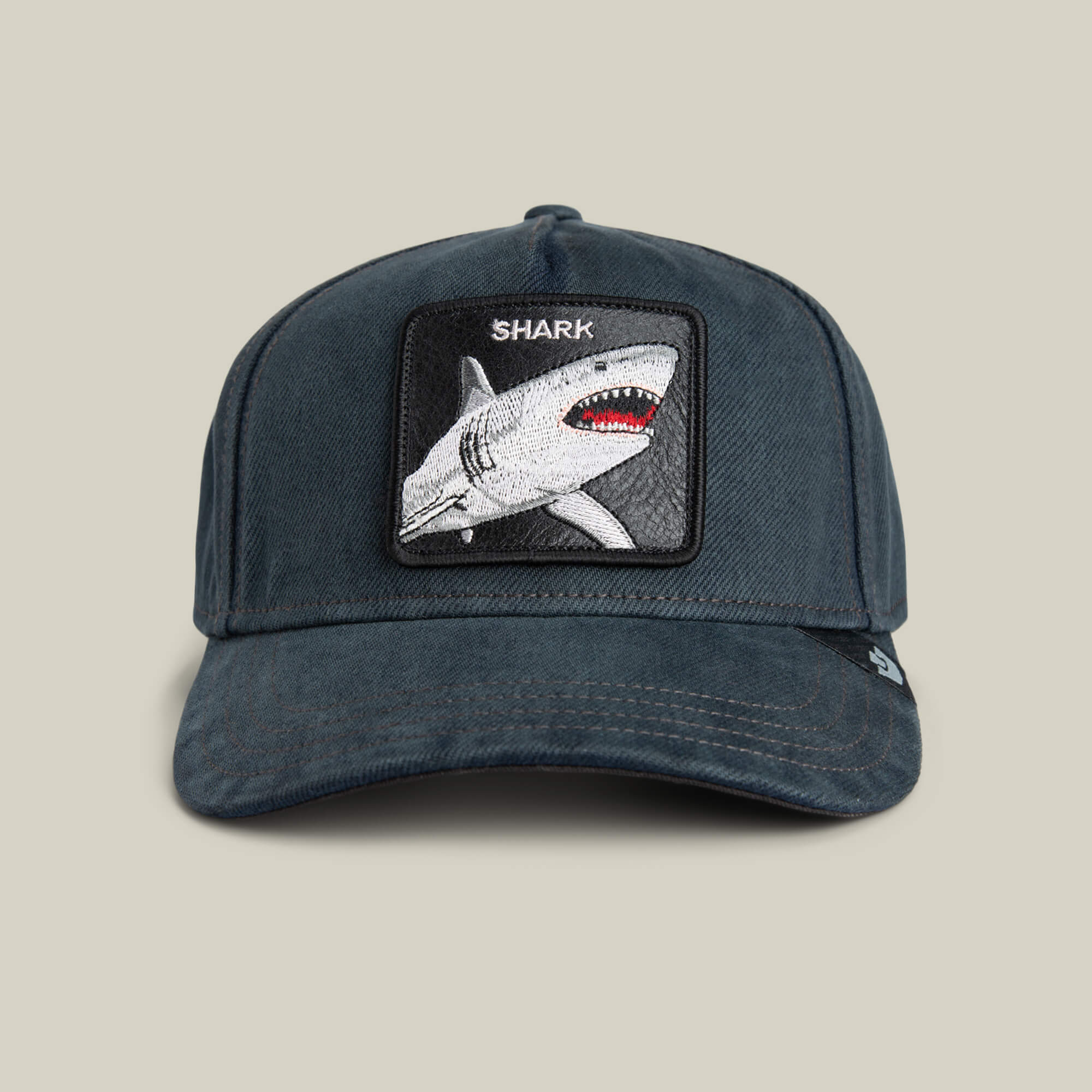 The "Smurf" by Goorin Bros. is a dark blue surf-punk cap made from overdyed denim, featuring a jagged grin patch with a shark image and "SHARK" embroidered above it.