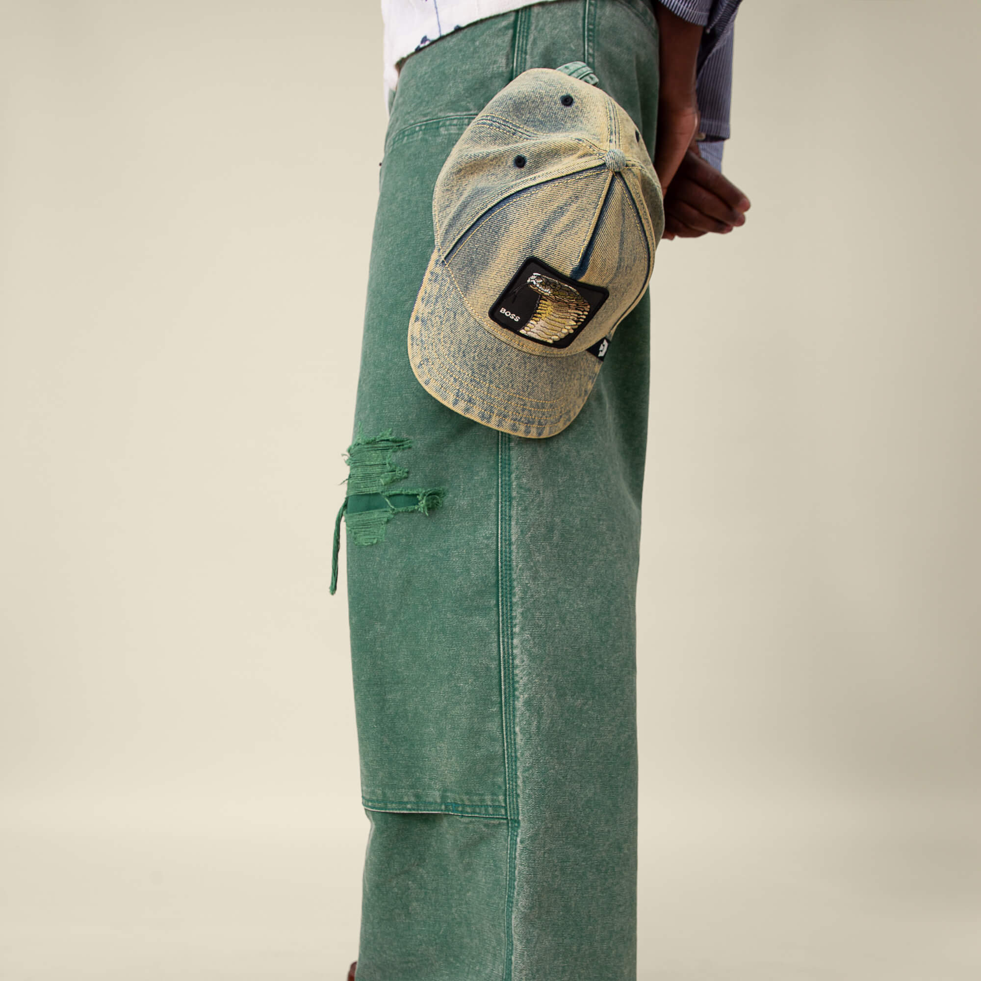 A person in loose green jeans with a ripped section holds the Goorin Bros. Pit Boss distressed denim hat, featuring a bold Cobra patch, shown from the waist down.