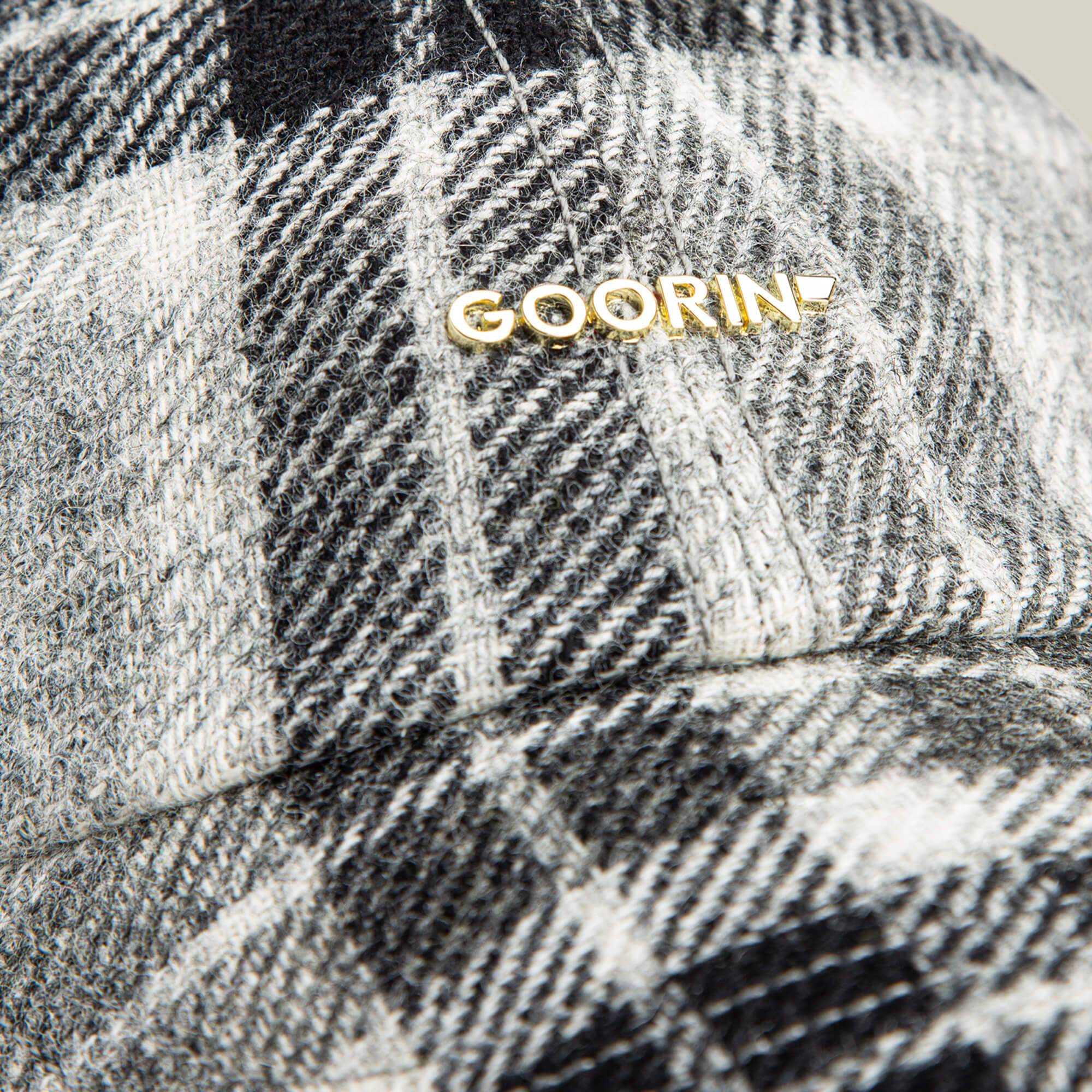 Close-up of the Goorin Bros. Bespoke Wordmark cap, a black, white, and gray plaid wool blend design with a gold Goorin metal logo near the seam.