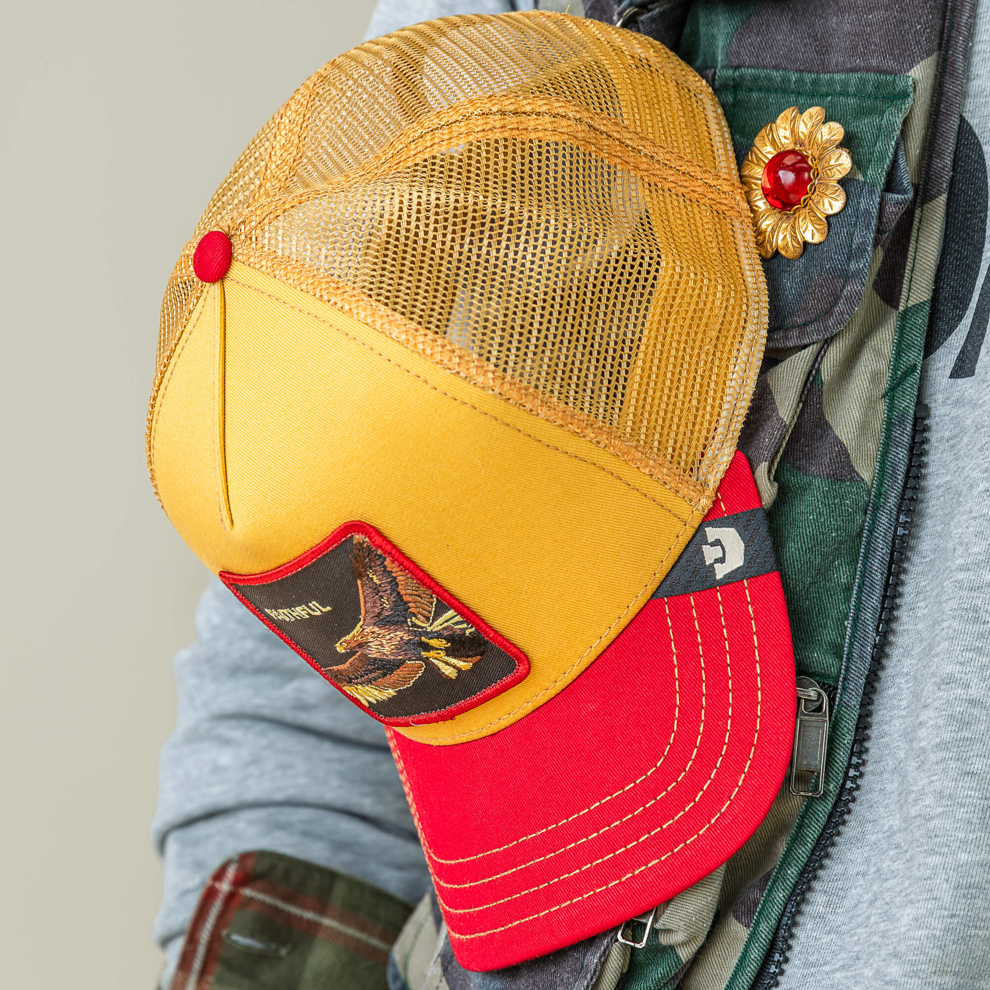The Faithful Eagle cap by Goorin Bros., featuring a yellow and red mesh design and bird patch, is paired with a camouflage jacket accented by a gold brooch set with a red gem.