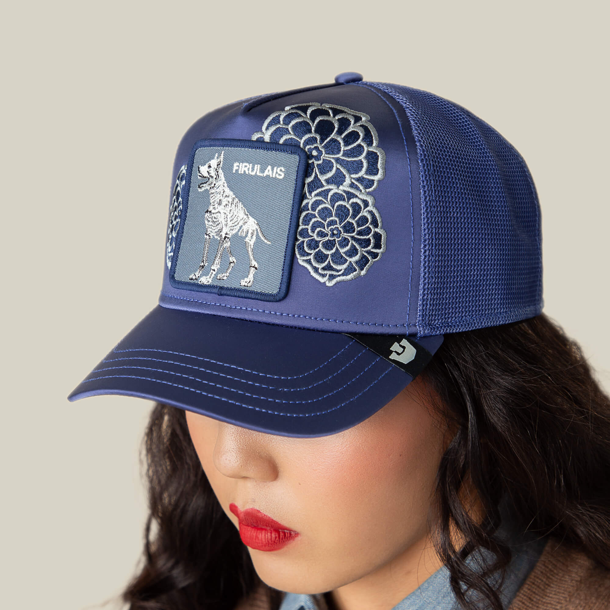 Person wears a blue mesh baseball cap by Goorin Bros., called Dia De Firulais, featuring embroidered floral patterns and a Firulais patch, celebrating Life After Ordinary.