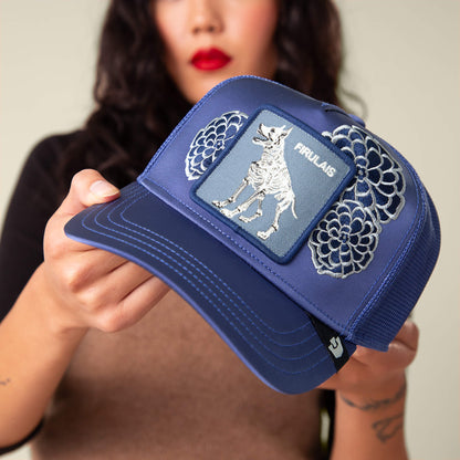 A woman holds a blue mesh trucker hat from Goorin Bros., called Dia De Firulais, featuring a mythical creature patch, floral embroidery, and the slogan "Life After Ordinary.