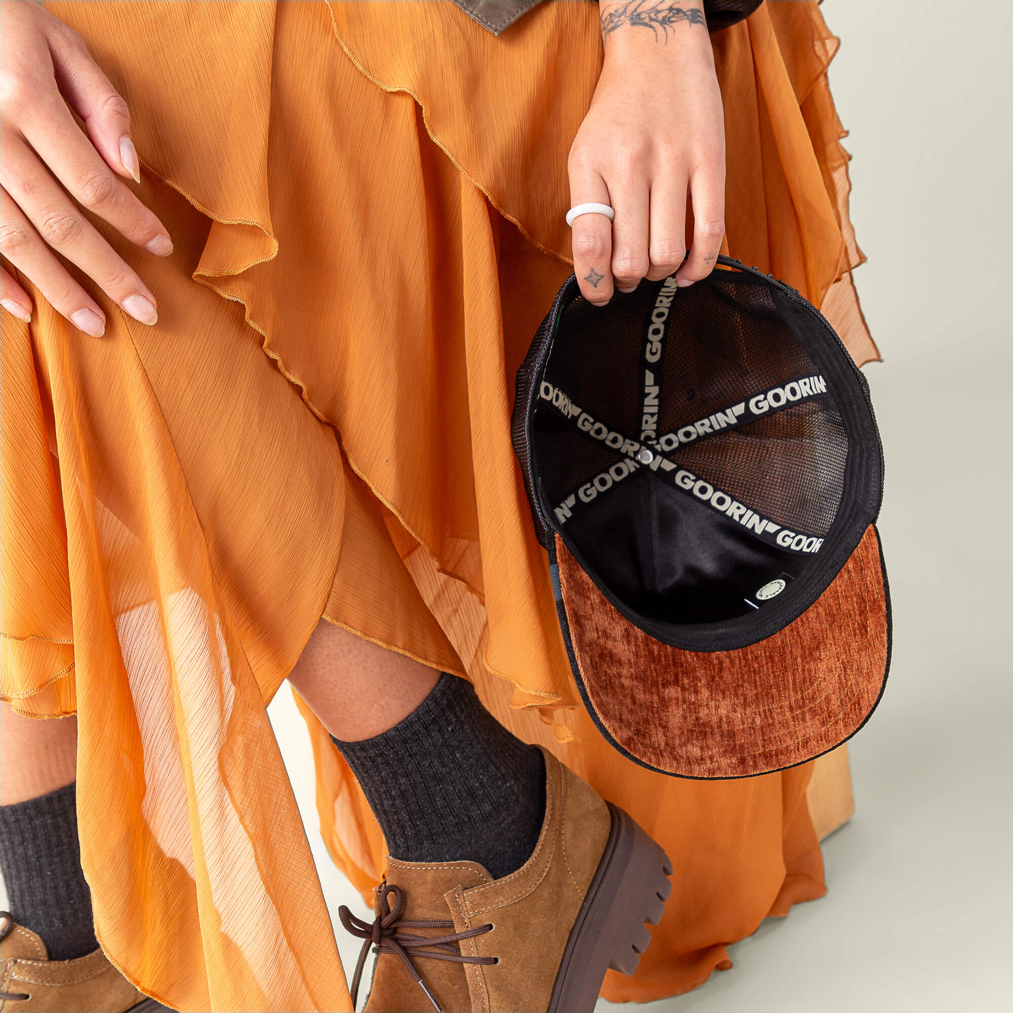 A person in a pleated orange skirt and brown shoes holds an open Goorin Bros. "Iced Panther" cap, revealing the signature interior branding—a hint of midnight velvet style.