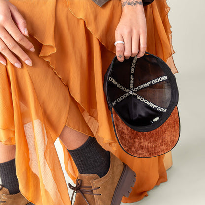 A person in a pleated orange skirt and brown shoes holds an open Goorin Bros. "Iced Panther" cap, revealing the signature interior branding—a hint of midnight velvet style.