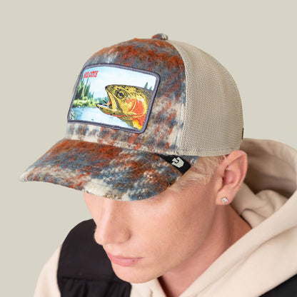A person wearing the Goorin Bros. "Down by the River" multicolored mesh trucker hat with a fish patch, paired with a tan hoodie, black vest, and flannel details—perfect for your next fishing trip.