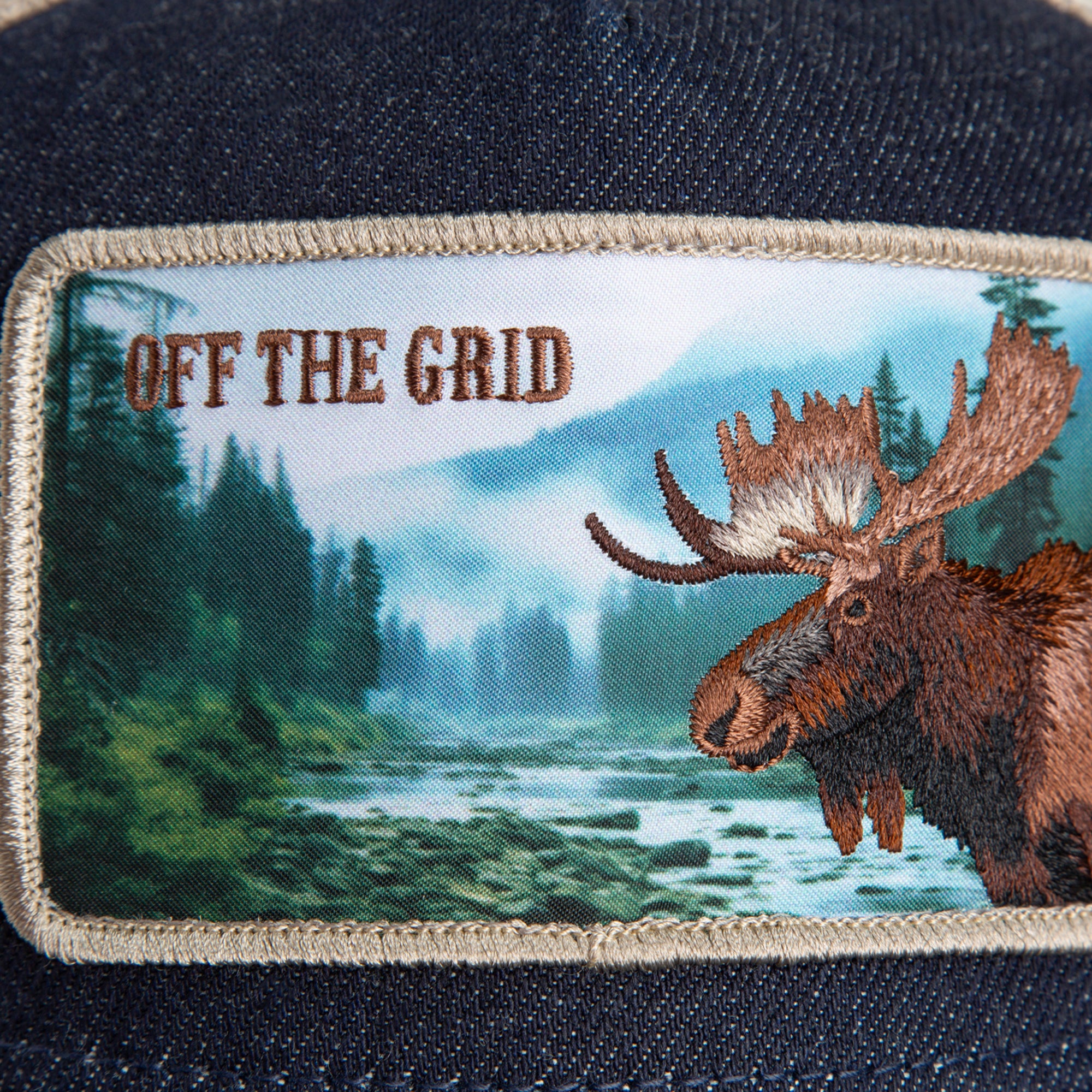 The Staycation patch by Goorin Bros. features a moose on denim with a forest river and mountains in the background, plus "OFF THE GRID" in bold brown capitals.