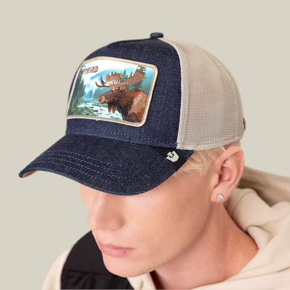 A person wearing the Goorin Bros. "Staycation" blue and beige mesh trucker hat, featuring a moose patch with a wilderness scene and mountains on the front.