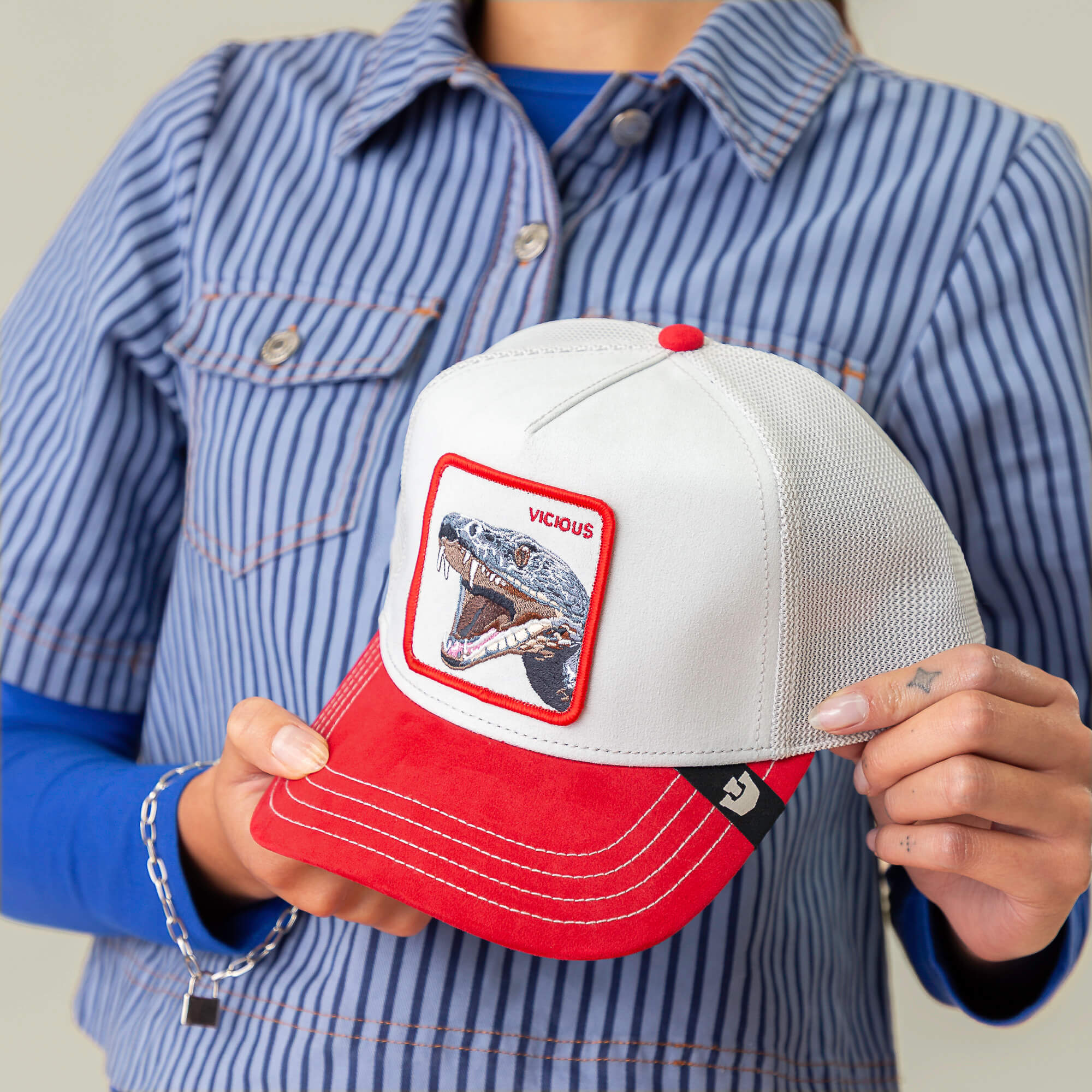 A person in a blue striped shirt holds the Goorin Bros. Suede Snake trucker cap in white and red, featuring a snarling snake patch with “VICIOUS” above it—radiating true predator energy.