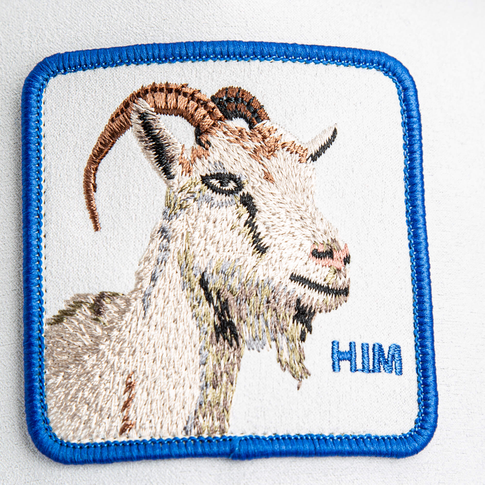 The Goorin Bros. Suede Goat patch features a brown and cream embroidered goat, blue outline, and "HIM" stitched in electric blue—ideal for adding standout style to your white crown hat.