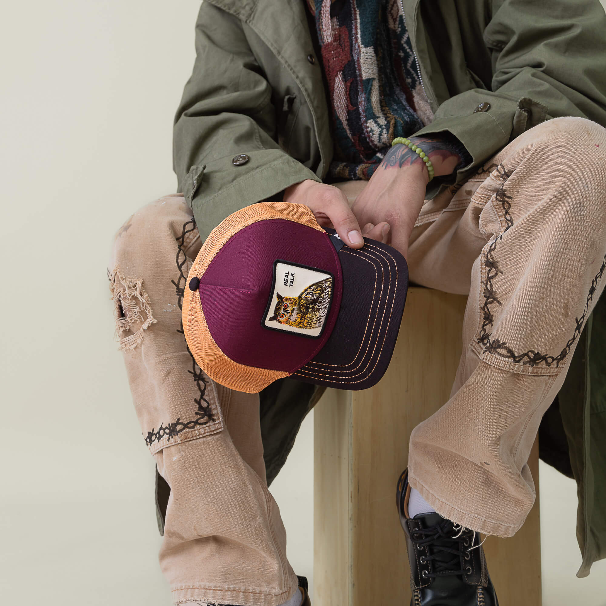 A person sits on a wooden stool, holding the Goorin Bros. Tricolor Owl black brim cap with an owl patch, paired with distressed jeans, black boots, and an olive green coat.