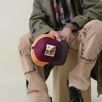 A person sits on a wooden stool, holding the Goorin Bros. Tricolor Owl black brim cap with an owl patch, paired with distressed jeans, black boots, and an olive green coat.