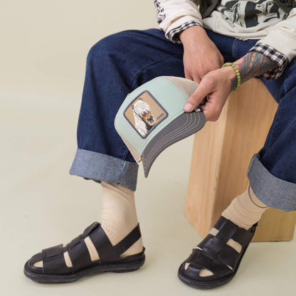 Seated on a wooden stool, a person wears cuffed jeans, socks, and black sandals while holding the Goorin Bros. Tricolor Horse hat featuring an animal patch.