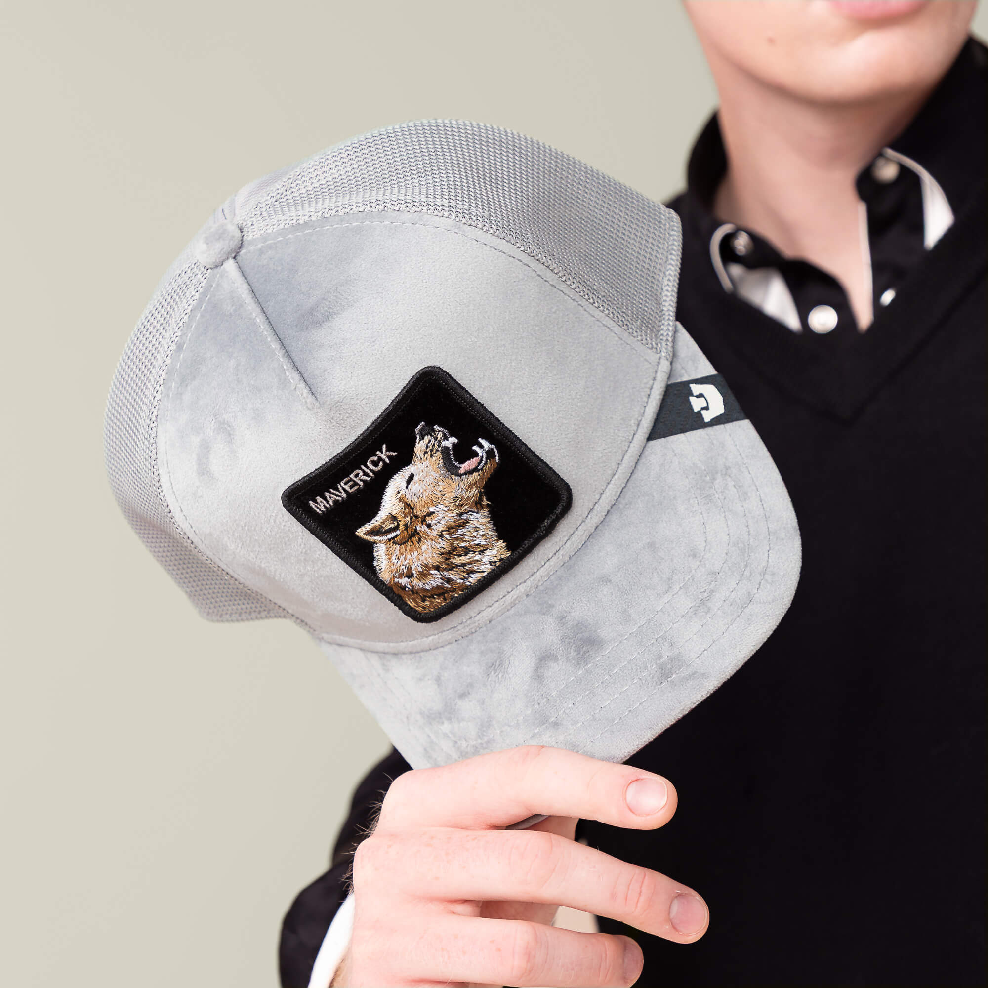 A person in a black sweater and striped-collar shirt holds the light-gray Velour Wolf cap by Goorin Bros., featuring a wolf patch—ideal for relaxing in the lounge or heading to your main event.