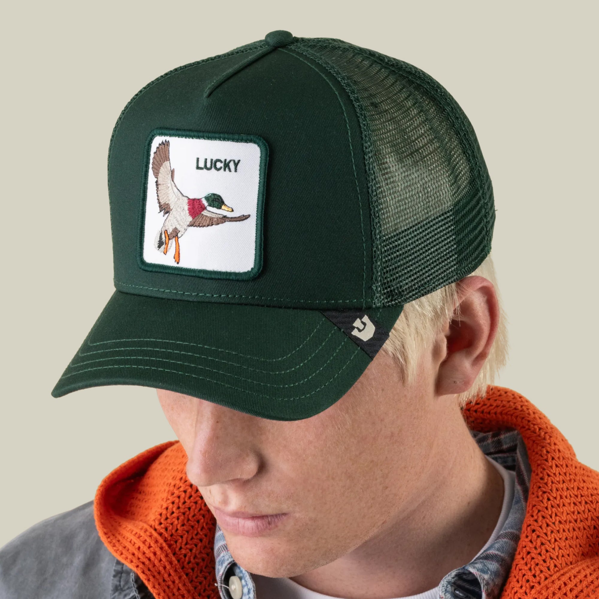 A person wearing Goorin Bros.’s The Lucky Duck dark green mesh trucker hat, featuring a flying duck fortune bird luck patch with the word “LUCKY,” along with an orange hoodie and denim jacket.
